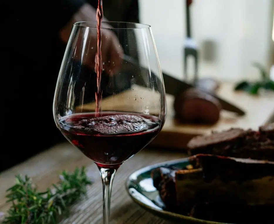 Nahaufnahme von Weinglas, in welches Rotwein eingeschenkt wird, im Hintergrund wird Fleisch zugeschnitten